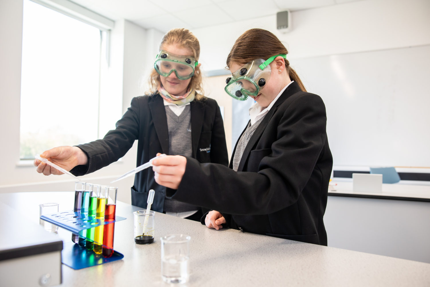 Two female students wearing safety goggles while using pipettes to drop in substances in a test tube