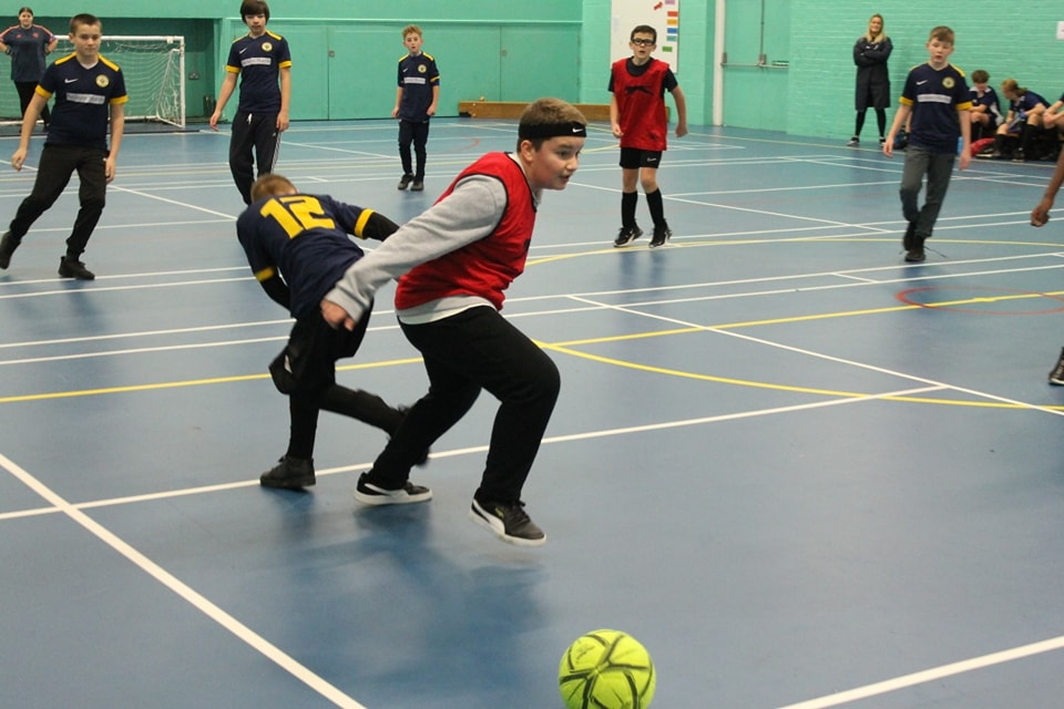 A group of boys are seen playing Football together in a Sports Hall, wearing PE Kit.