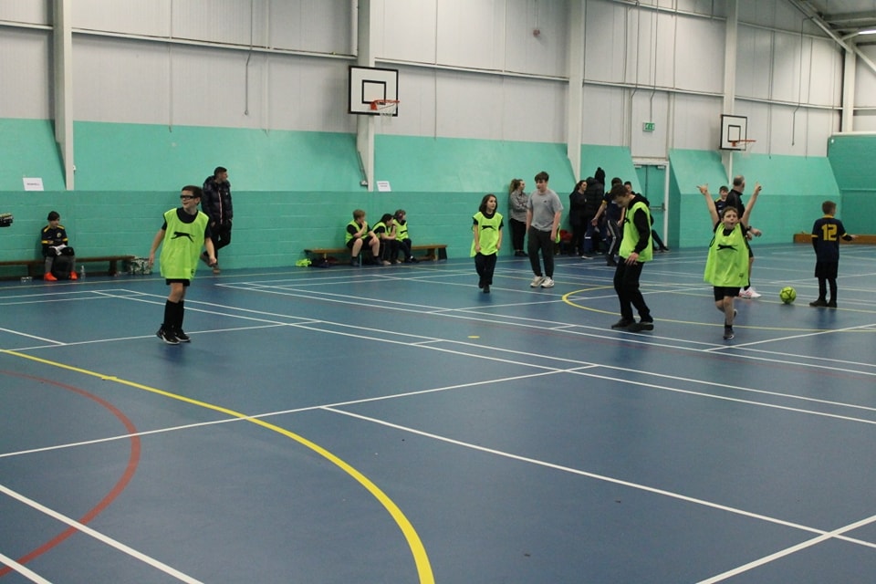 A group of Snowfields Academy students are shown wearing fluorescent bibs whilst playing a game of Football in the Sports Hall.
