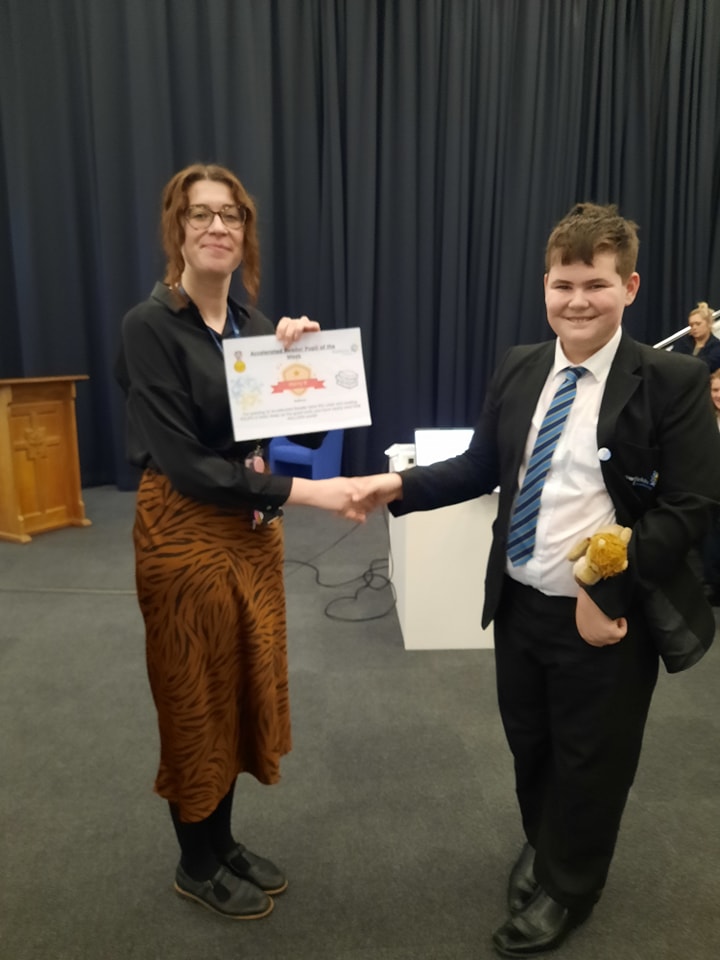 A student is seen posing for a photo for the camera, whilst collecting a certificate he has won from a female member of staff.