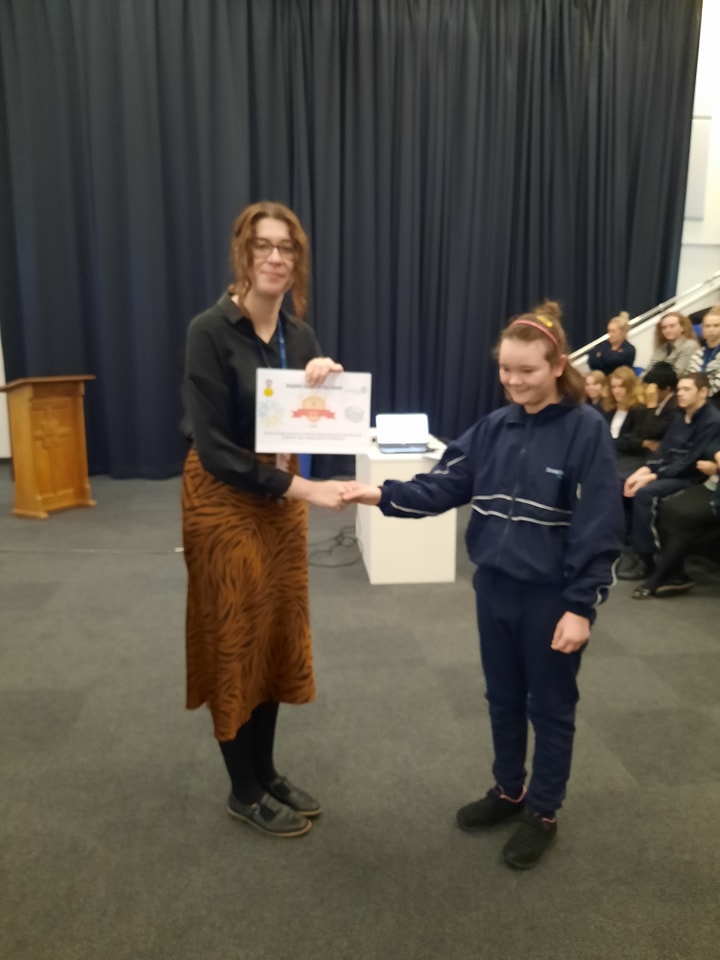A student is seen posing for a photo for the camera, whilst collecting a certificate she has won from a female member of staff.