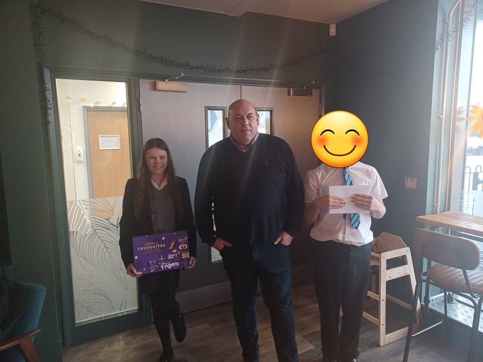 Two students are pictured smiling for the camera alongside an adult member of staff, holding prizes they have won for completing an activity.