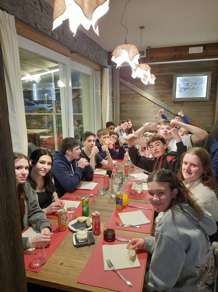 A large group of students from Snowfields Academy can be seen sat around a long table, as they prepare to have dinner during a skiing trip abroad.